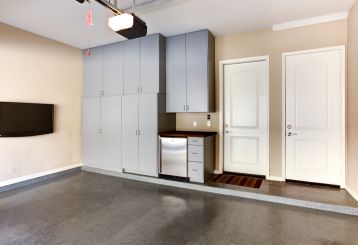 Modern garage interior with sleek gray storage cabinets, wall-mounted TV, mini fridge, and two white entry doors in Blaine, Minnesota.