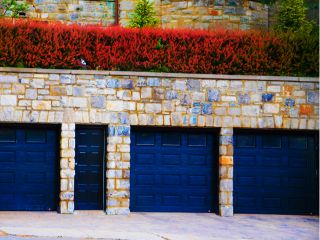 Row of three black garage doors set in a stone wall with red shrubs above, representing well-maintained garage doors in Blaine, Minnesota. Three Black Garage Doors with Stone Exterior in Blaine MN